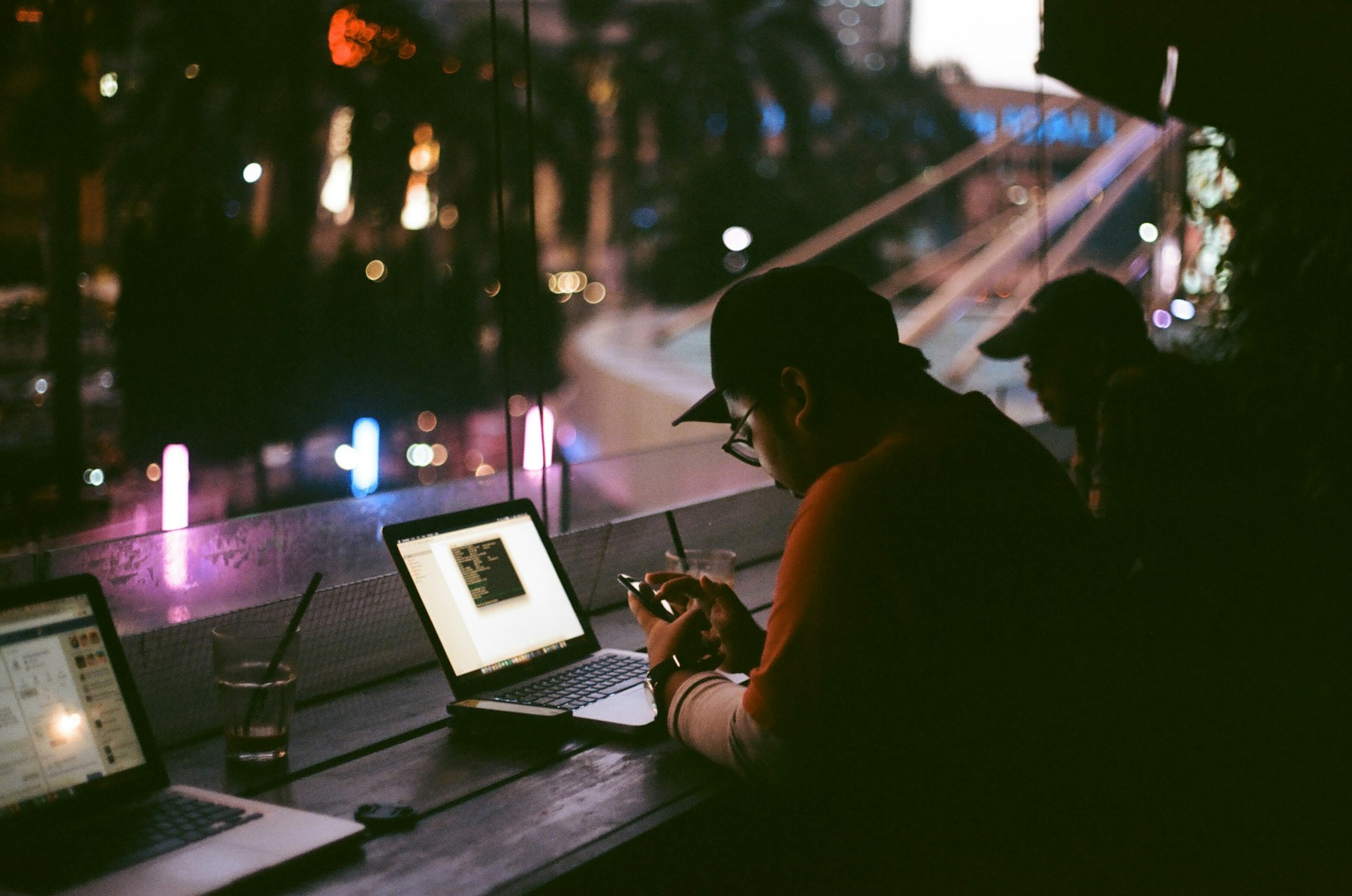 A person working alone at a laptop at night, with a phone in hand and a city visible through the window